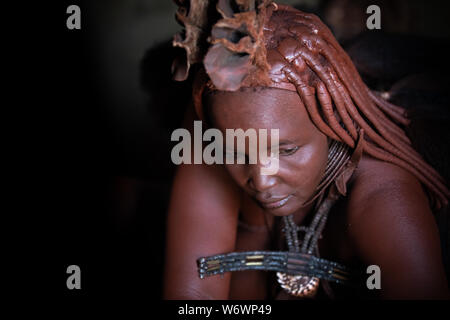 Opuwe, Namibia - Juli 12, 2019: Ein traditionelles Himba Frau in der Dunkelheit ihrer Lehmhütte. Stockfoto