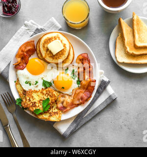 Amerikanisches Frühstück auf grau Tabelle, aus der Nähe. Sonnenseite gebratene Eier, gebratenen Speck, Hash Brown, Pfannkuchen, Orangensaft und Kaffee zum Frühstück. Stockfoto