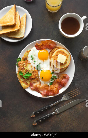 Amerikanisches Frühstück auf dunklen, Ansicht von oben, kopieren. Sonnenseite gebratene Eier, gebratenen Speck, Hash Brown, Pfannkuchen, Toast, Orangensaft und Kaffee für Stockfoto