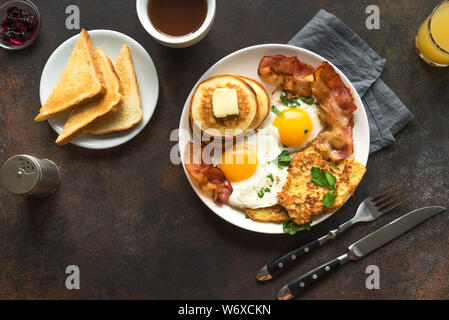 Amerikanisches Frühstück auf dunklen, Ansicht von oben. Sonnenseite gebratene Eier, gebratenen Speck, Hash Brown, Pfannkuchen, Toast, Orangensaft und Kaffee zum Frühstück. Stockfoto