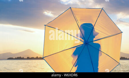 Silhouette der junge Hipster in Hut mit Regenschirm. Kind mit Regenschirm suchen im Seascape. Reisen Konzept. Sommerferien Hintergrund. Auf Wiedersehen im Sommer. Abschied vom Sommer. Inkognito Hintergrund Stockfoto