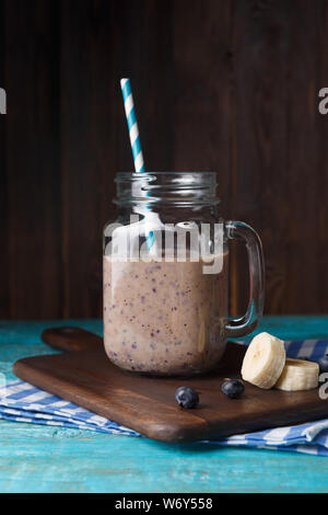 Foto von Kanne mit Milch Cocktail mit Stroh, Banane und Heidelbeeren auf schneidebrett an der hölzernen Hintergrund Stockfoto