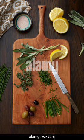 Kräuter, Oliven und Zitronen auf einem Holzschneidebrett mit Messer, Serviette und Salzschüssel auf einem Holztisch Stockfoto