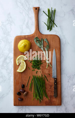 Kräuter, Oliven und Zitronen auf einem Holzschneidebrett mit einem Messer auf einer Marmoroberfläche Stockfoto