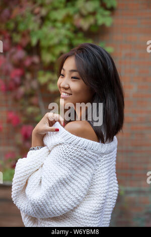 Schöne asiatische Mädchen mit einem Lächeln auf ihrem Gesicht in Weiß Strickjacke in der Nähe von Mauer und lockig wild bunte Trauben Stockfoto