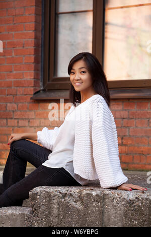 Schöne glücklich lächelnde asiatischen Mädchen sitzt auf einem Stein Treppe in einem weißen Strickjacke in der Nähe einer Mauer auf der Straße Stockfoto