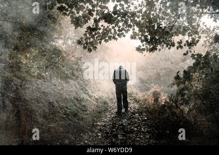 Eine vermummte Gestalt zu Fuß entfernt von der Kamera an einem nebligen Wald Weg mit den Schultern und blickte nach unten. Mit einem Grunge, Vintage bearbeiten Stockfoto