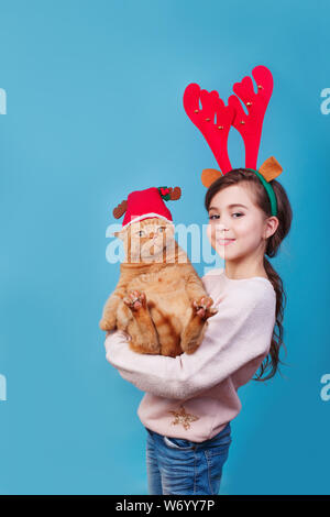 Lustige kleine lächelnde Mädchen mit roter Katze in Rentiergeweih auf blauem Hintergrund. Frohe Weihnachten und Frohes neues Jahr. Stockfoto