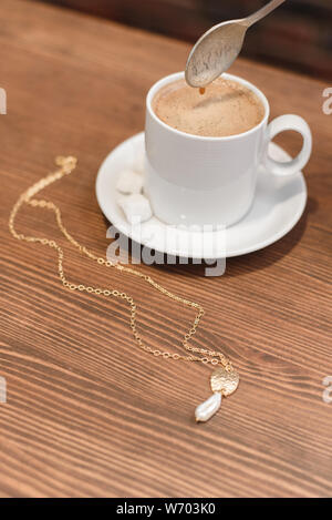 Schmuck auf dem Hintergrund von frisch gebrühtem Kaffee auf einem Holztisch. Mädchen rührt Kaffee mit Milch. Gold Schmuck mit Perlen auf dem Tisch. Stockfoto