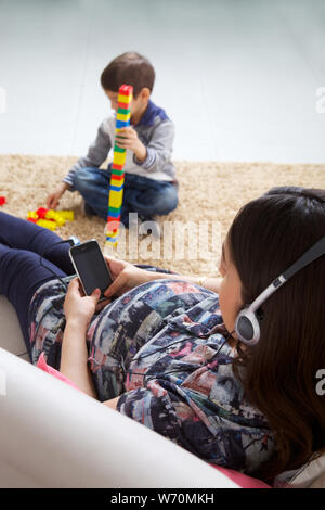 Schwangere Mutter anhören von Musik auf einem Mobiltelefon mit ihrem Sohn spielen Stockfoto