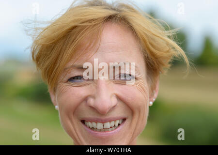 Glückliche Frau mit einer warmen, freundlichen Lächeln und ordentlich kurzen roten blondes Haar in einer Nahaufnahme Kopf Porträt im Freien Stockfoto