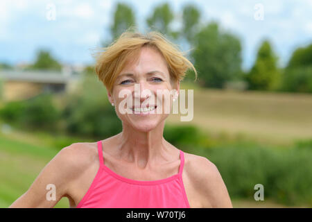 Verspielt blonde Frau, eine käsige toothy Grinsen, als sie sich mit den Händen auf den Hüften draußen im Land Stockfoto