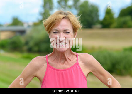 Verspielt blonde Frau, eine käsige toothy Grinsen, als sie sich mit den Händen auf den Hüften draußen im Land Stockfoto