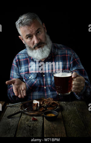 Brutale grauhaariger erwachsenen Mann mit einem Bart Senf Steak isst und trinkt Bier, lädt ein zu einem Essen, das Konzept eines Urlaubs, Festival, Oktoberfest oder St. Pa Stockfoto