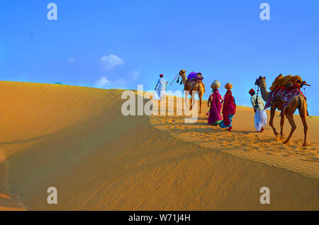 Zwei Kameltreiber und Frauen mit Kamelen Wandern auf Sanddünen der Wüste Thar gegen den blauen Himmel, Jaisalmer, Rajasthan, Indien Stockfoto
