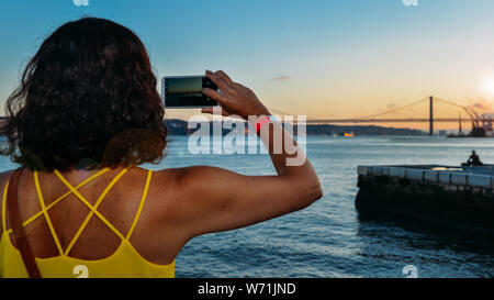 Unbekannte Frau bei Sonnenuntergang erfassen Sie ein Bild auf Ihr Handy von 25 de Abril Brücke über den Tejo in Lissabon, Portugal. Stockfoto