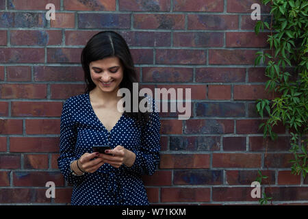 Bild der glückliche junge Frau gegen die Wand gelehnt und mit Smartphone Stockfoto