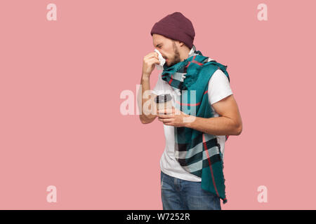 Portrait von Krankheit moderne bärtigen Jungen hipster Mann im weißen Hemd und casual Hut stehend, Niest der Serviette, Holding heißen Getränk in Pappbecher. Innen- Stockfoto