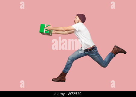 Seitenansicht des Portrait von Crazy bärtigen Jungen hipster Mann im weißen Hemd und casual Hut springen, laufen und sich beeilen mit der Lieferung grün präsentieren. Indo Stockfoto