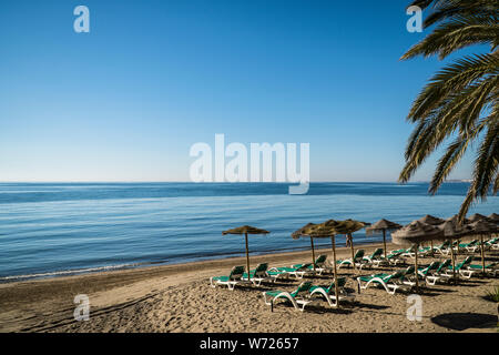 Blick auf den Strand von Marbella, Spanien. Mit leeren Liegestühlen, Stroh Schirme, Mittelmeer und strahlend blauer Himmel Stockfoto