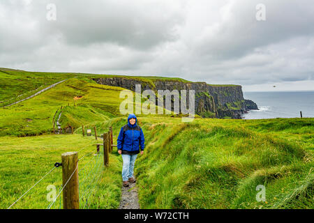 Frau in Blau Jacke zu Fuß der Küste zu Fuß auf dem Weg von Doolin in die Klippen von Moher, geosites und Geopark, wilden Atlantik Weg, Frühling regnerischen Tag Stockfoto