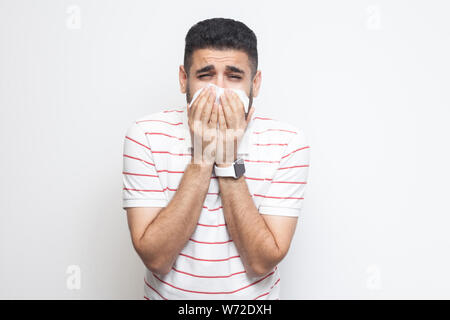 Erkältung und Grippe. Portrait von Kranken bärtiger junger Mann in gestreiften T-Shirt, die hielten Gewebe auf Mund und Kamera mit Traurigkeit und Krankheit. Stockfoto