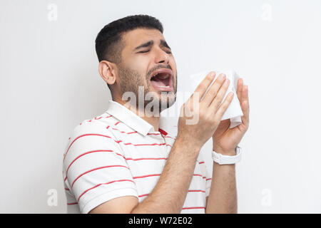 Erkältung und Grippe. Portrait von Kranken bärtiger junger Mann in gestreiften T-Shirt, die hielten Gewebe und Niesen. indoor Studio shot, isoliert auf weißem Hinterg Stockfoto