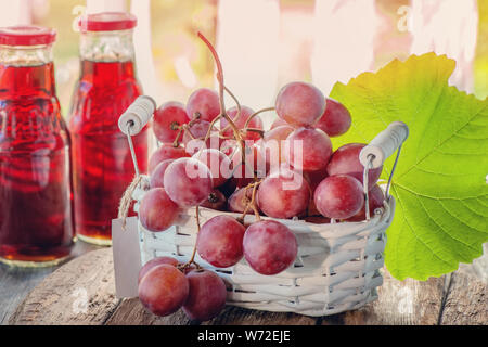 Ein Bündel rosa Trauben, vorbereitet, um den Saft zu extrahieren, ist in einem weißen Korb . Zwei Flaschen Traubensaft stehen auf dem Tisch neben einem Traubenhaufen Stockfoto