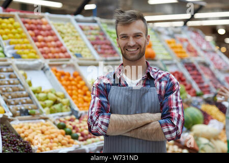 Taille bis Portrait von gutaussehenden jungen Mann im Supermarkt arbeiten und lächelnd an der Kamera, während durch Obststand Posing, kopieren Raum Stockfoto