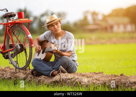 Asiatischer Mann Gitarre zu spielen, die grüne Reisfelder in den Sonnenuntergang. Auf dem Land von Thailand Stockfoto