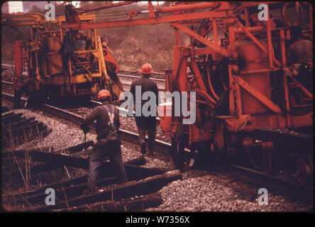 SOUTHERN RAILWAY VORFAHRT ARBEITER UND MASCHINEN. Die Maschine BUBEN DIE SCHIENEN, ZIEHT DIE ALTE CROSSTIES UND STELLEN NEUE, TEMPEL DIE BINDUNGEN AN, fährt dann Spikes auf den Schienen in Position zu halten. Wendete das Unternehmen 18,6 Prozent des Umsatzes 1974 auf die Wartung der Gleisanlagen. In diesem Jahr SOUTHERN HATTE EINEN DURCHSCHNITT VON VIER Mannschaften von 50 bis 55 Männer jedes ARBEITEN AN DER ANLAGE 10,531 Meilen Schiene Stockfoto