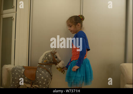 Kleines Mädchen spielt mit einem Pferd Gurney. Das Kind sitzt auf einem Pferd. Schöne Mädchen ist glücklich sitzen auf einem Spielzeug Pferd. Einen blauen Rock, eine orange Stockfoto