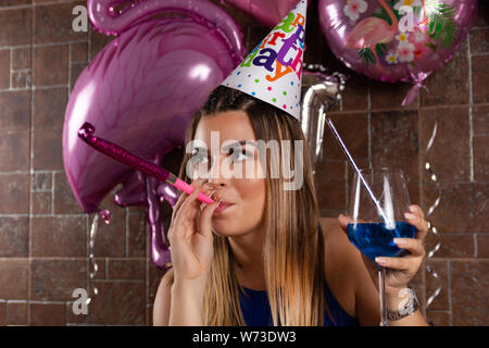 Glückliche junge Frau mit musikalischen Ausblasen und Cocktail Blue Lagoon in der Hand und die Kappe auf dem Kopf feiert ihren Geburtstag in einen Nachtclub. Geburtstag Stockfoto