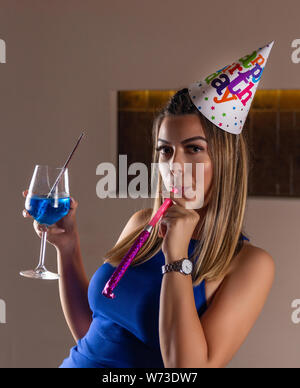 Attraktives Mädchen mit musikalischen Ausblasen und Cocktail Blue Lagoon in der Hand und die Kappe auf dem Kopf feiert ihren Geburtstag in einen Nachtclub. Geburtstag Stockfoto