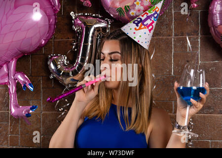 Gerne schöne Mädchen mit musikalischen Ausblasen und Cocktail Blue Lagoon in der Hand und die Kappe auf dem Kopf feiert ihren Geburtstag in einem Nachtclub Stockfoto