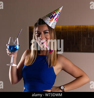 Glückliches Mädchen mit musikalischen Ausblasen und Cocktail Blue Lagoon in der Hand und die Kappe auf dem Kopf feiert ihren Geburtstag in einen Nachtclub. Geburtstag Konzept Stockfoto