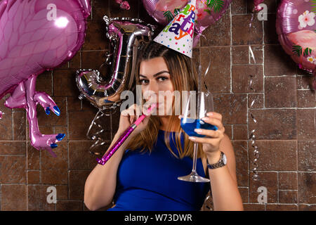 Glückliche junge Mädchen mit musikalischen Ausblasen und Cocktail Blue Lagoon in der Hand und die Kappe auf dem Kopf feiert ihren Geburtstag in einen Nachtclub. Geburtstag Stockfoto