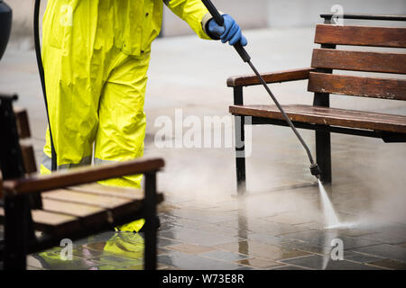Bürgersteig und Bänke in einer Einkaufsstraße, Jet gewaschen und von einem Arbeiter Gelb hohe vis Kleidung sprühen Wasser aus einem Schlauch gereinigt Stockfoto