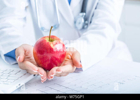 Frau Doktor im Krankenwagen hält roten Apfel in die Hände. Auf dem Schreibtisch Laptop und EKG-Diagramm. Stockfoto