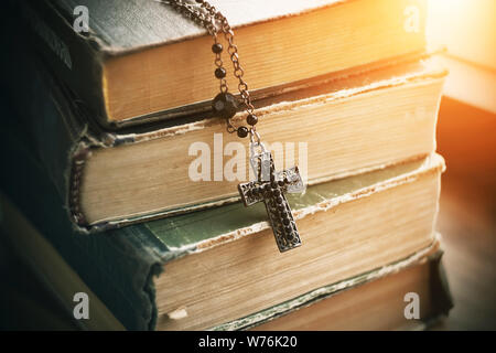 Auf alten dicken Bücher über Schrift und Magie liegt ein schwarzes Metall Kreuz auf eine Kette mit Perlen, durch ein helles Licht beleuchtet. Stockfoto