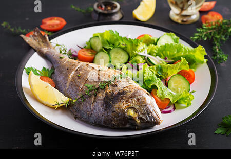 Gebackenen Fisch dorado mit Zitrone und frischen Salat in weiße Platte auf dunklen rustikalen Hintergrund. Gesund essen mit Fisch Konzept. Diät und sauber Essen Stockfoto