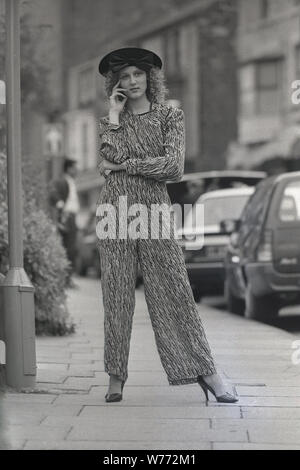 1980er Jahre, draußen auf einem Bürgersteig stehend, posierte eine attraktive junge Frau in der neuesten weiblichen Mode, ein Stylist-Hosenanzug, auch bekannt als Jumpsuit, England, Großbritannien. Stockfoto