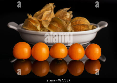 Menge ganze Frische orange Physalis in weiße, ovale Keramik Schüssel in der Zeile auf schwarzem Glas isoliert Stockfoto