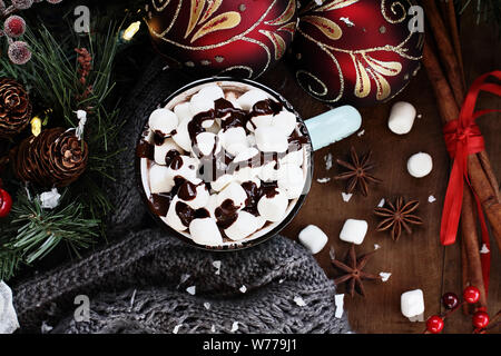 Emaille Tasse heißen Kakao zu Weihnachten mit Mini Marshmallows und garniert mit Schokoladensauce.  Umgeben von warmen grauen Schal, Ornamente, Kiefer Ast Stockfoto