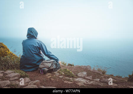 Ansicht der Rückseite des Wanderer Frau sitzt auf der Kante einer Klippe mit blauen Regenmantel und Rucksack am Horizont und Meer. Stockfoto