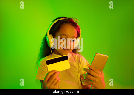 Schöne weibliche Brustbild auf grünem Hintergrund im Neonlicht isoliert. Junge emotionale Mädchen. Menschliche Gefühle, Mimik Konzept. Mit Smartphone für online Zahlungen Rechnung. Stockfoto