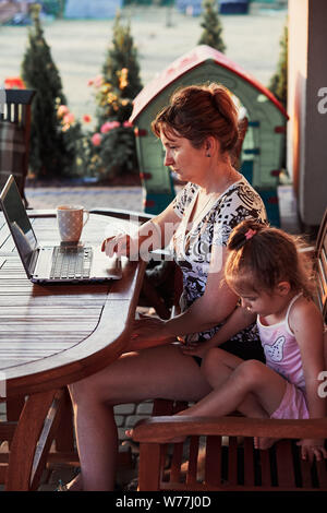 Mutter zu Hause arbeiten, mit tragbaren Computer, sitzen auf der Terrasse mit ihrer Tochter im Sommer Tag Stockfoto