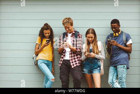 college-Freunde, die Taschen tragen, indem sie Mobiltelefone im Freien benutzen. Multiethnische Teenager-Jungen und -Mädchen stehen draußen an einer Wand und schauen sich an Stockfoto