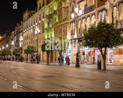 Eine breite Straße mit tramtracks, Geschäfte und Menschen waling in der Nacht in der Altstadt von Sevilla, Spanien Stockfoto