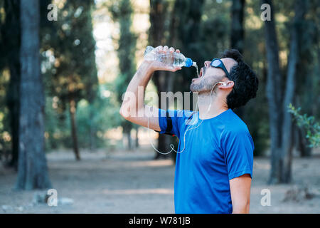 Weißer Mann Trinkwasser und das Hören von Musik in einem Park während des Trainings. Stockfoto